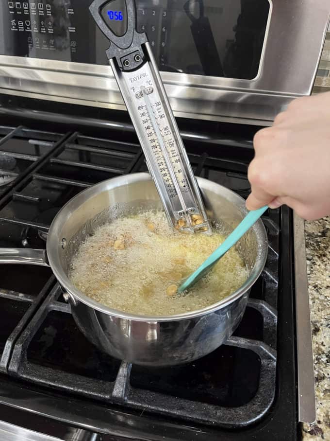 a pot on a stovetop with granulated sugar, water, corn syrup and peanuts cooking in it being mixed with a rubber spatula and a candy thermometer attached to the side