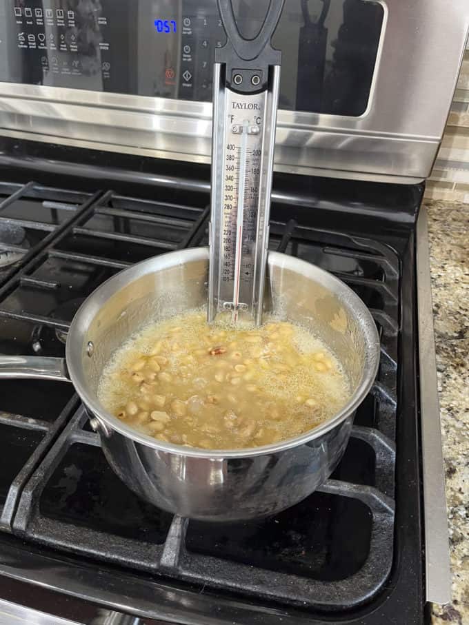 a pot on a stovetop with granulated sugar, water, corn syrup and peanuts cooking in it and a candy thermometer attached to the side