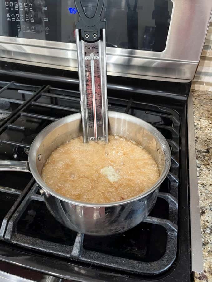 a pot on a stovetop with granulated sugar, water, corn syrup, baking soda, vanilla extract and peanuts cooking in it and a candy thermometer attached to the side