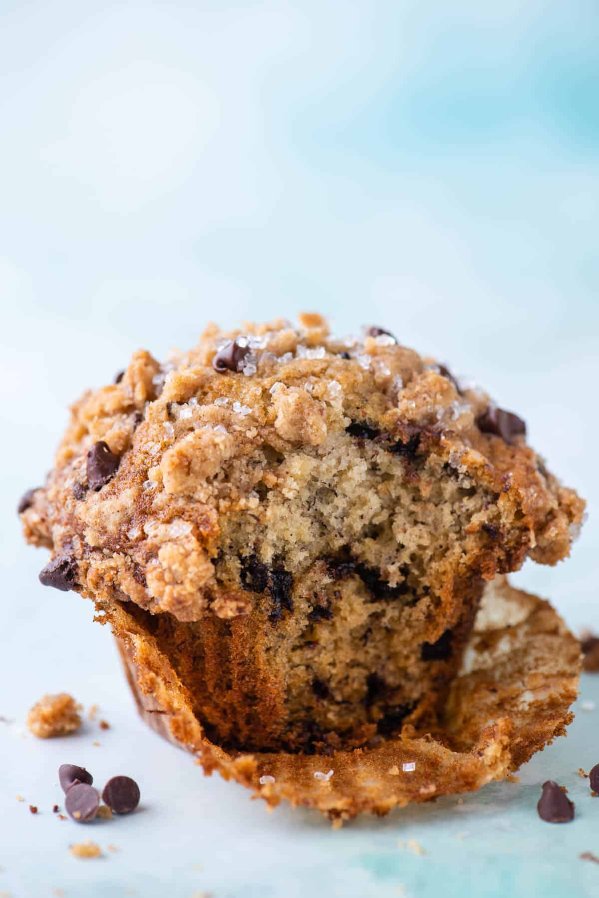 a banana chocolate chip muffin sitting on a light teal surface with its muffin liner peeled partially open, a bite taken out of the muffin, surrounded by muffin crumbs and mini chocolate chips