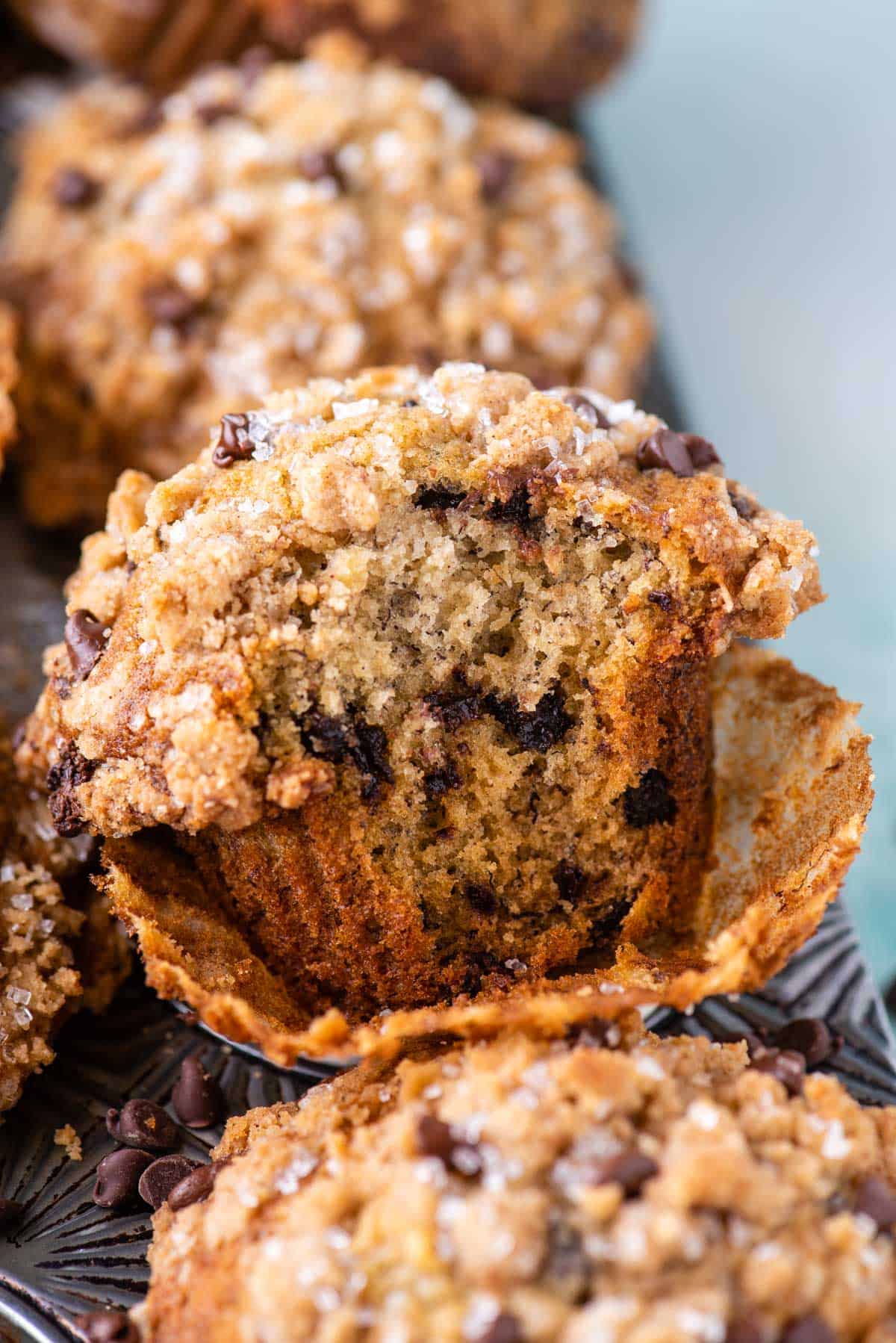a banana chocolate chip muffin with its muffin liner pulled open and a bite missing, sitting on top of a muffin pan full of more muffins