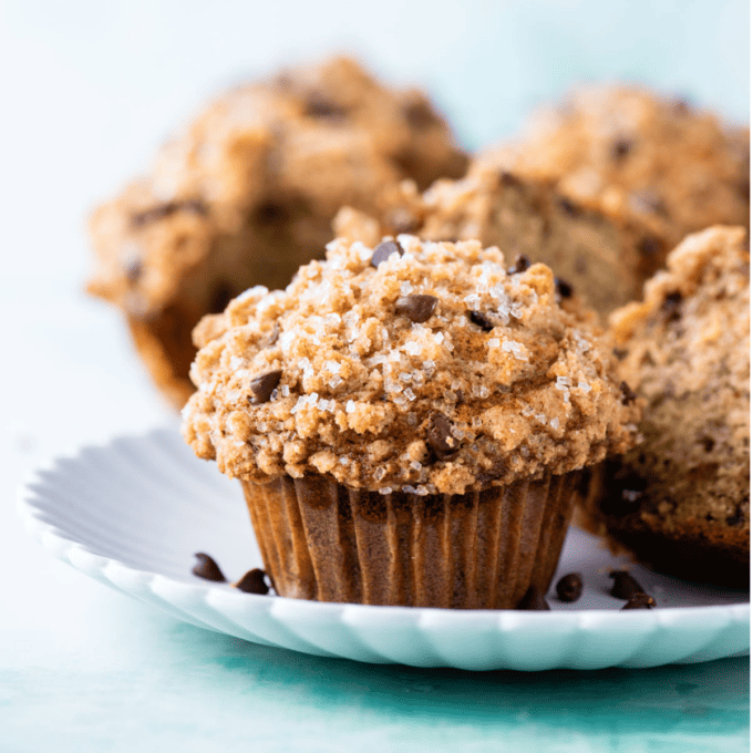 banana chocolate chip muffins on a white plate sitting on a light teal surface with chocolate chips sprinkled around