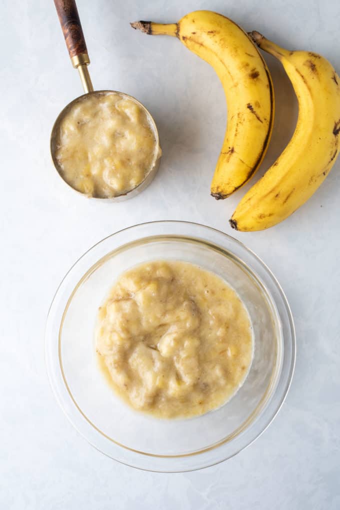 a clear glass bowl full of mashed banana beside a measuring cup full of mashed banana and two whole bananas