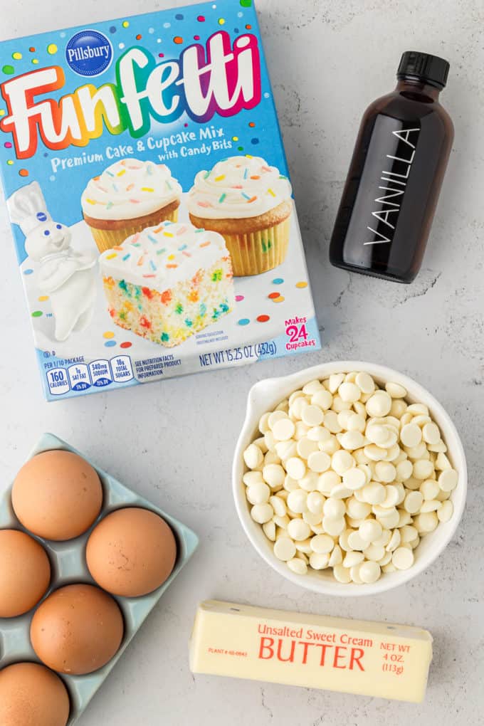 a box of funfetti cake mix, a bottle of vanilla extract, a bowl of white chocolate chips, a crate of eggs, and a stick of butter