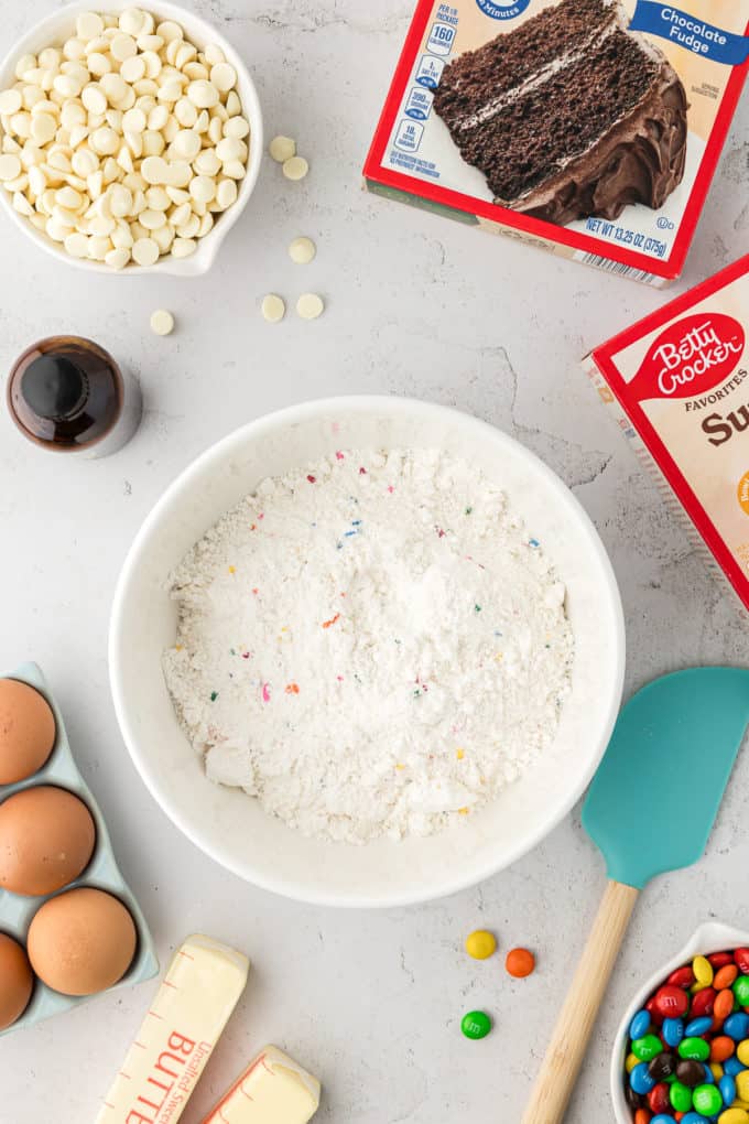 a bowl of cake mix in the middle with boxes of cake mix, a bowl of white chocolate chips, a bottle of vanilla, eggs, butter, a rubber spatula and a bowl of M&Ms surrounding it.