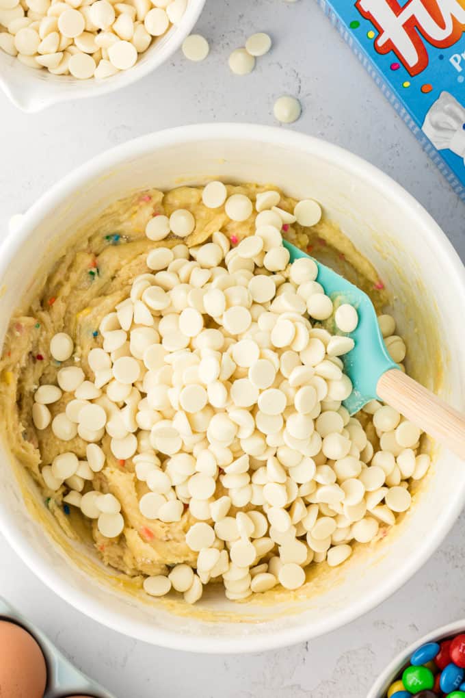 a white bowl full of funfetti cake bar mix with a rubber spatula in it and a pile of white chocolate chips on top, a cake mix box, eggs, a bowl of M&Ms, and a bowl of white chocolate chips surrounding the bowl