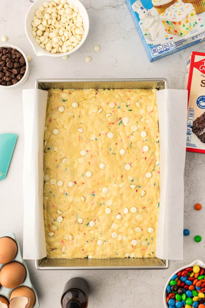 a rectangular pan of funfetti cake bar batter surrounded by two boxes of cake mix, eggs, a bottle of vanilla, a bowl of M&Ms, a small bowl of chocolate chips and a larger bowl of white chocolate chips