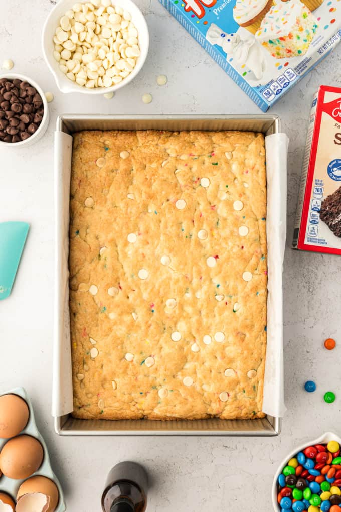 a rectangular pan of funfetti cake bars that haven't been cut yet, surrounded by two boxes of cake mix, eggs, a bottle of vanilla, a bowl of M&Ms, a small bowl of chocolate chips and a larger bowl of white chocolate chips