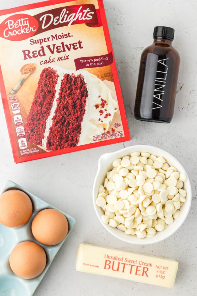 a box of red velvet cake mix, a bottle of vanilla extract, a bowl of white chocolate chips, a crate of eggs, and a stick of butter