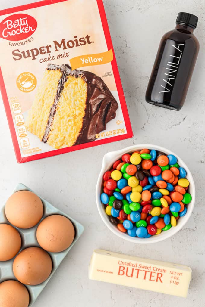 a box of cake mix, a bottle of vanilla extract, a bowl of M&Ms, a crate of eggs, and a stick of butter