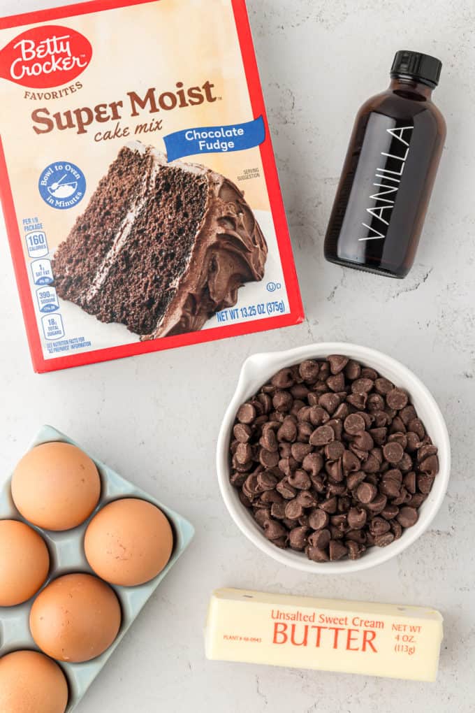 a box of chocolate cake mix, a bottle of vanilla extract, a bowl of chocolate chips, a crate of eggs, and a stick of butter