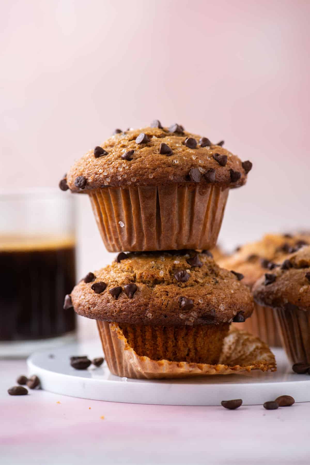 a stack of two cappuccino chip muffins with coffee beans sprinkled around, more muffins and a cup of coffee in the background