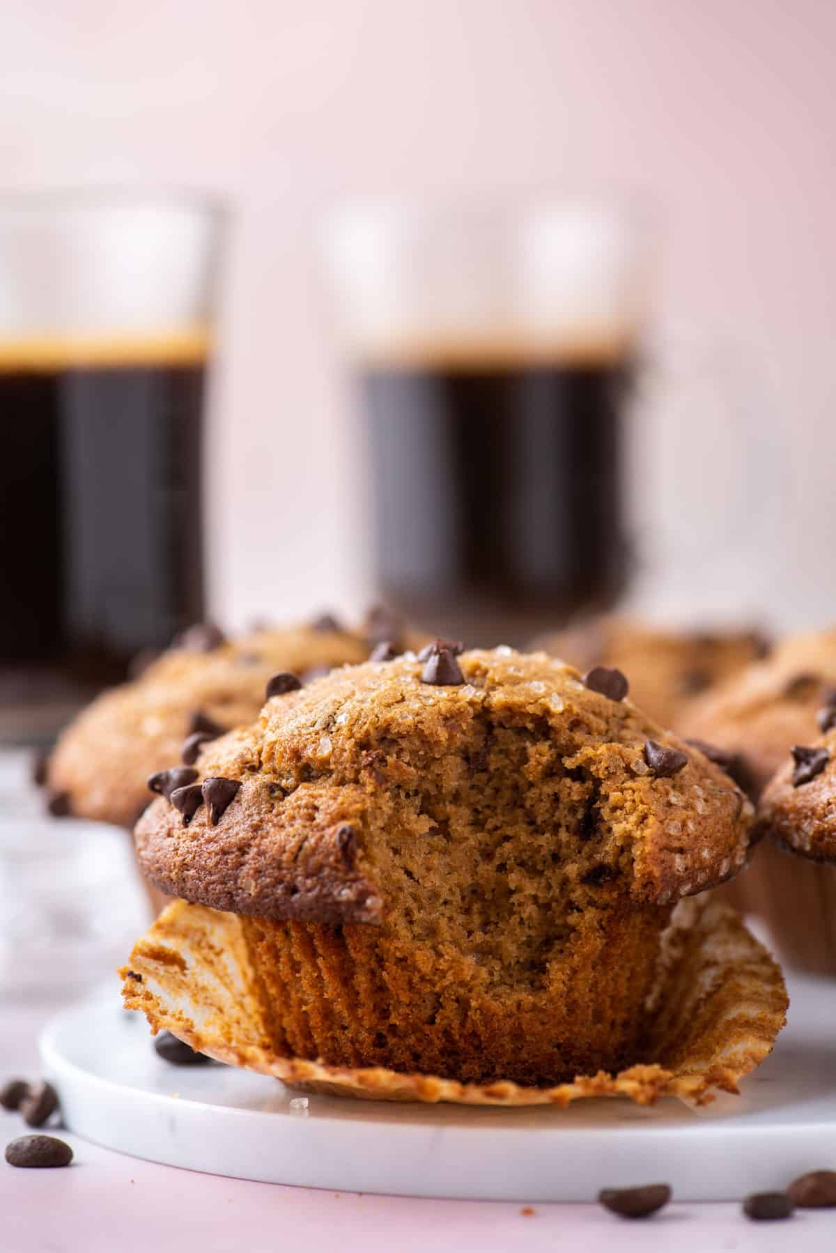 a cappuccino chip muffin on a white plate with a bite taken out, sitting in its open muffin liner, with coffee beans sprinkled around, more muffins and two cups of coffee in the background