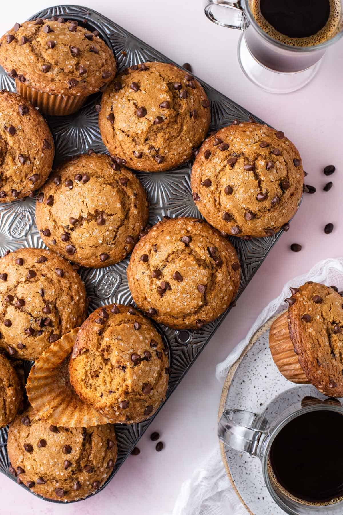 overhead view of a muffin pan full of cappuccino chip muffins with coffee beans sprinkled around, with a cup of coffee and a plate with a muffin and cup of coffee beside it