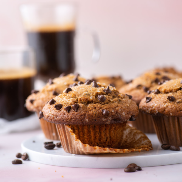 cappuccino chip muffins on a white plate with chocolate chips sprinkled around and two cups of cappucinno in the background