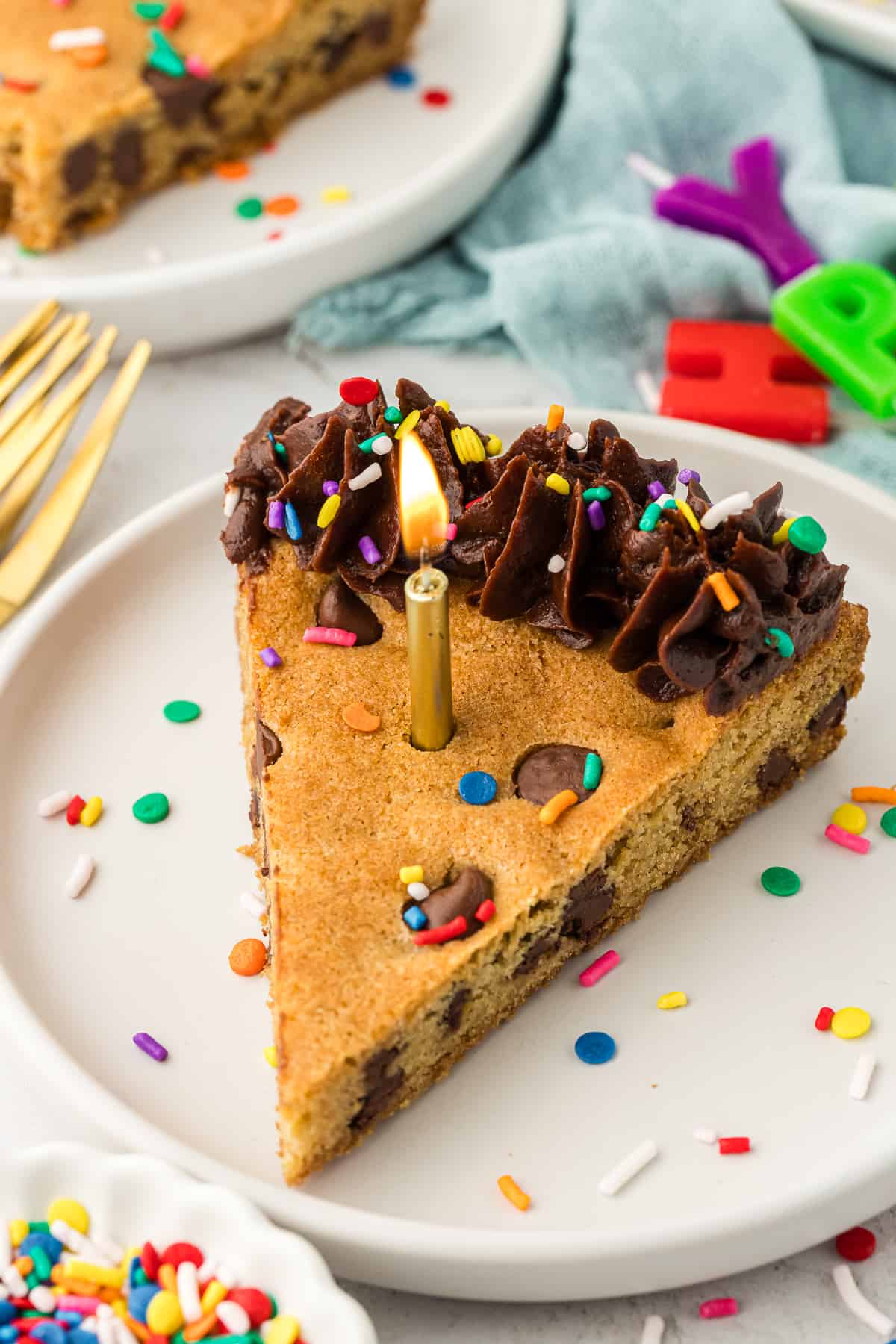 a slice of cookie cake on a white plate with a gold lit candle in the center of it, surrounded by gold forks, happy birthday letter candles and more plates with slices of cake