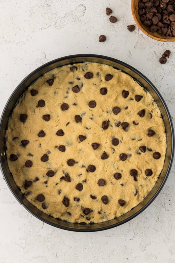 cookie cake batter in a round cake pan with a bowl of chocolate chips beside it
