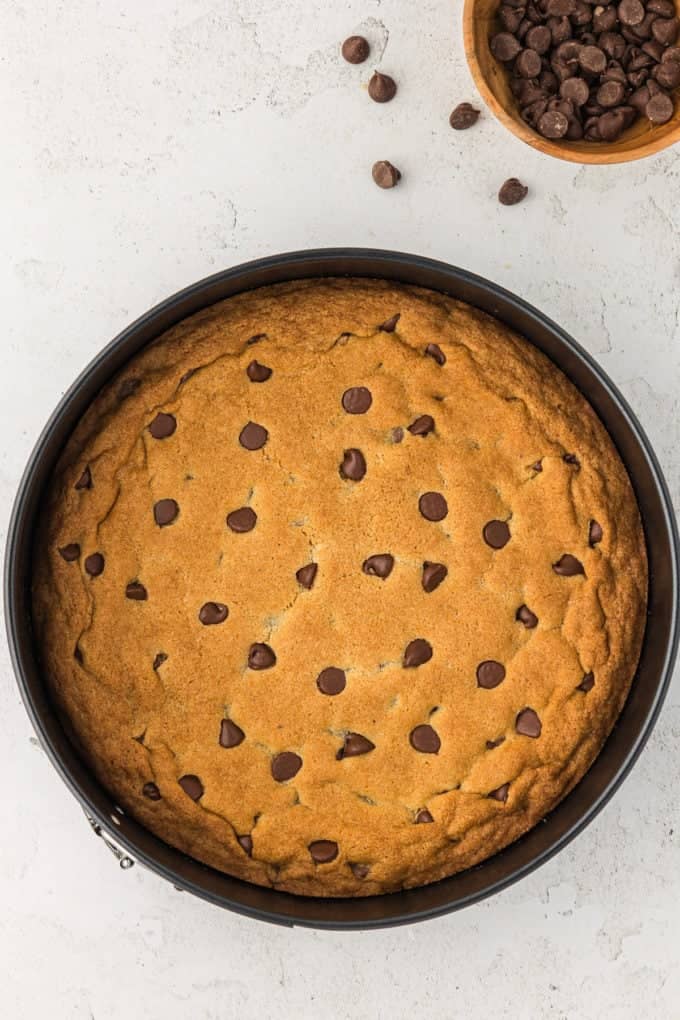 a freshly baked cookie cake in a round pan with a bowl of chocolate chips beside it