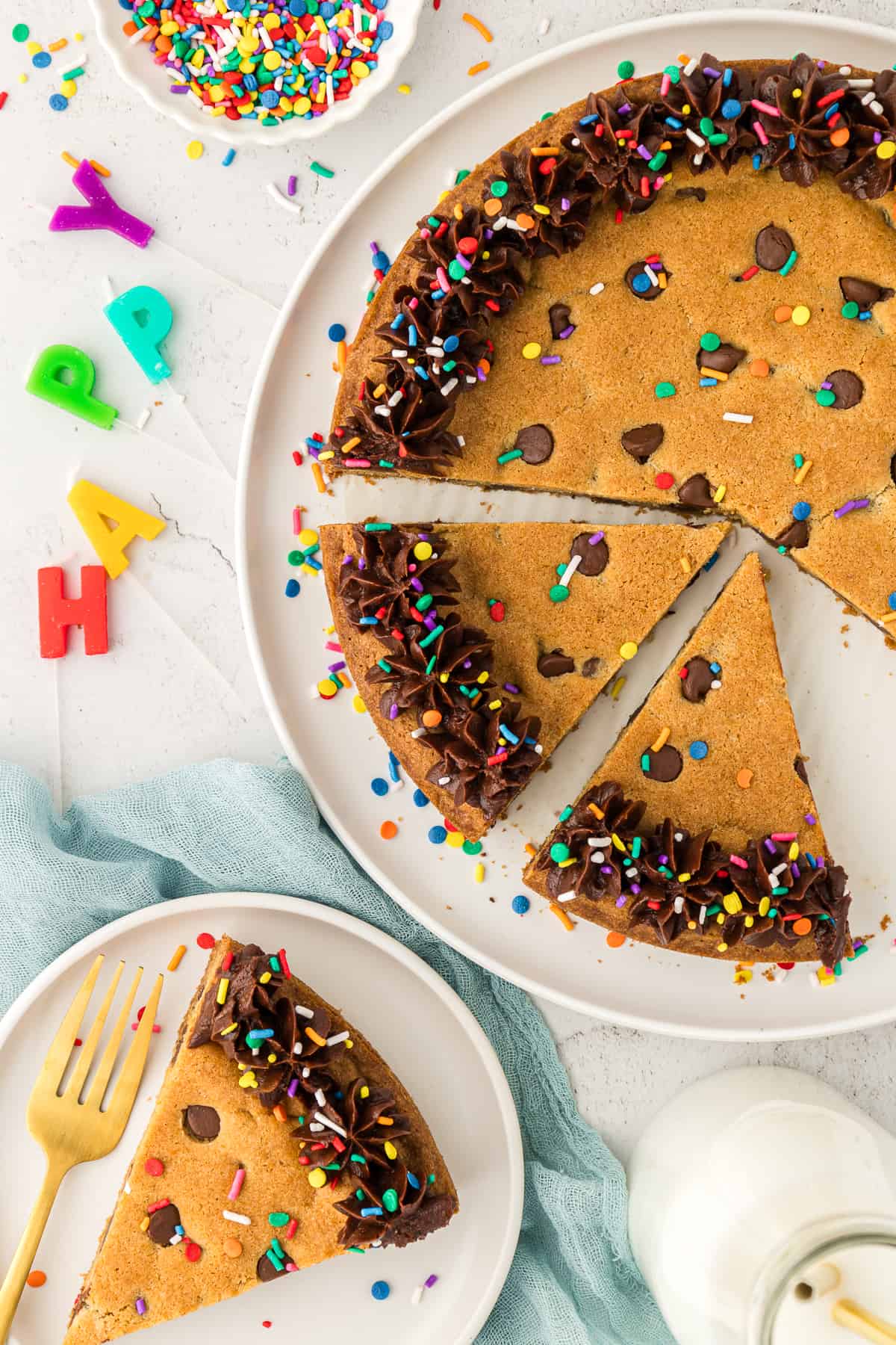 a cookie cake on a white plate with two slices cut, another slice on a smaller white plate with a form and happy birthday candles and a bowl of sprinkles beside it