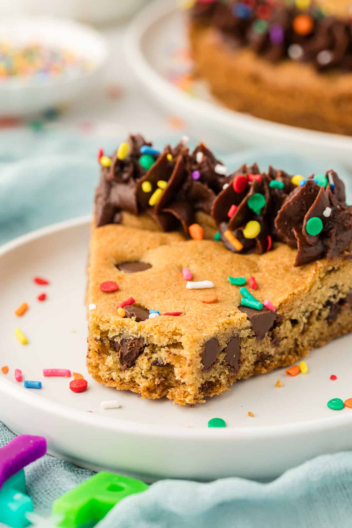 a slice of cookie cake on a white plate decorated with chocolate frosting and colored sprinkles, with one bite missing
