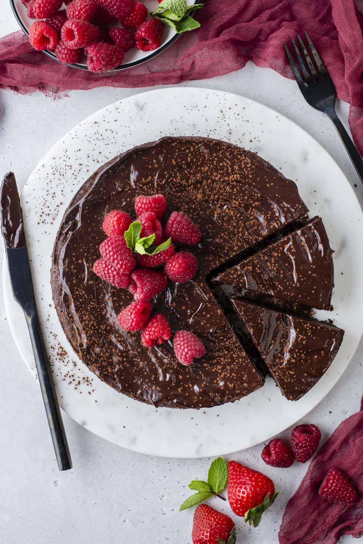 overhead shot of a full flourless chocolate cake with two slices cut, sitting on a round white serving platter, topped with fresh raspberries and mint leaves, with a knife covered in chocolate laying beside it, red clothes and more raspberries and fresh strawberries surrounding it