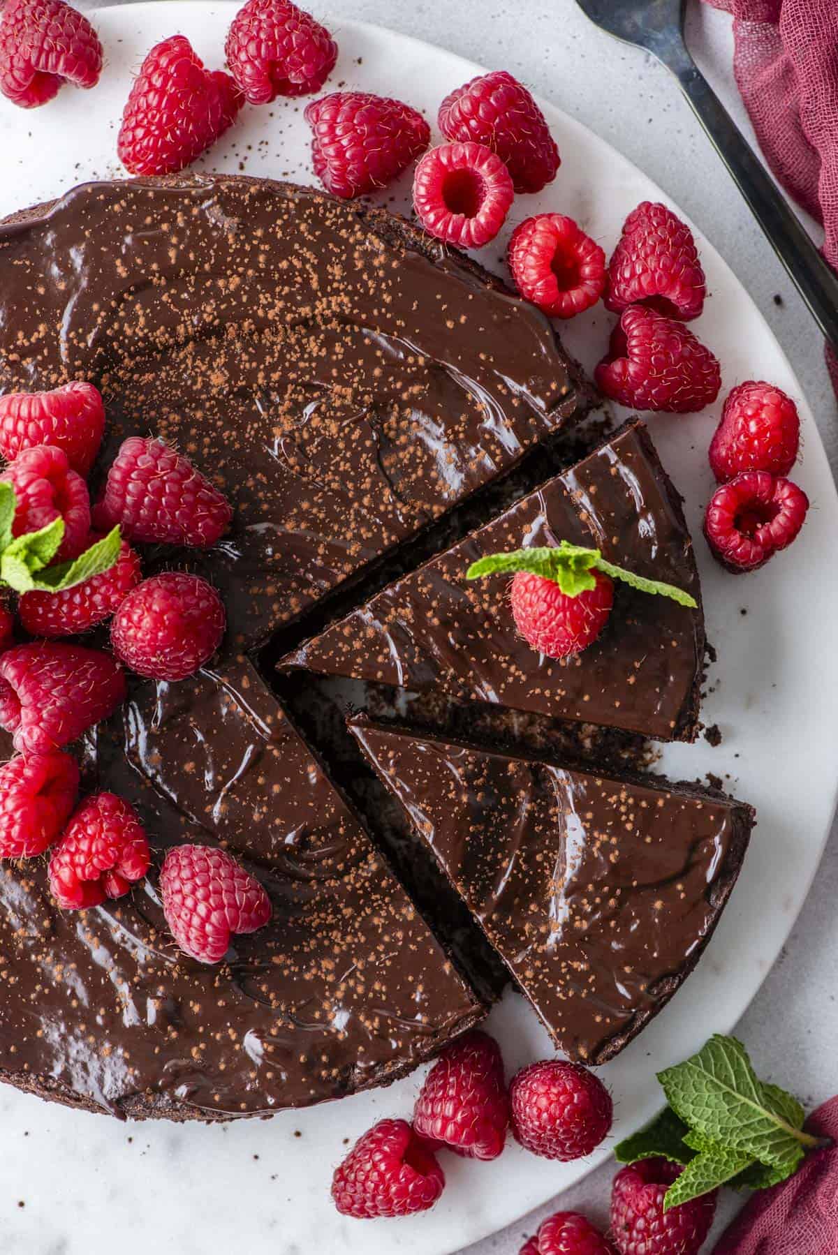 overhead view of a flourless chocolate cake with two slices cut, sitting on a white plate and topped with fresh raspberries and mint leaves