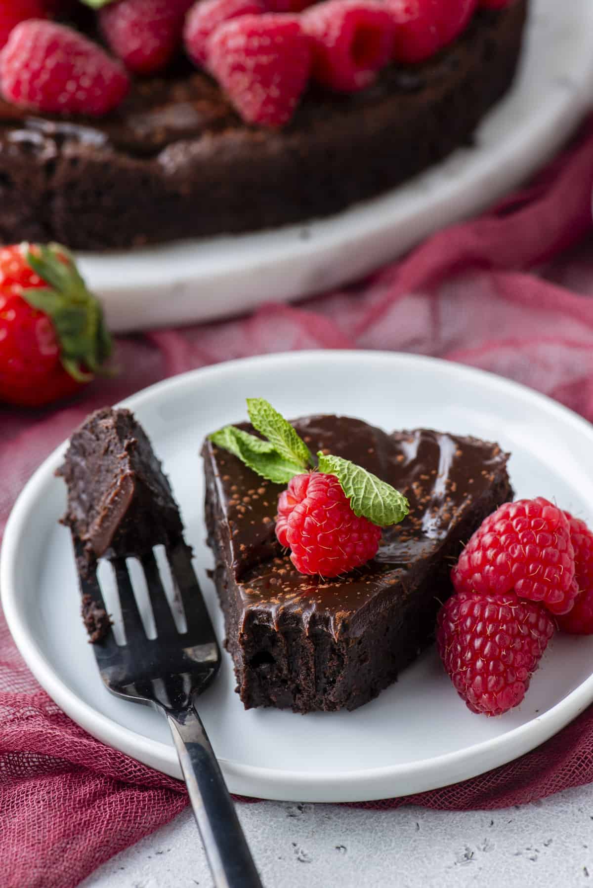 a slice of flourless chocolate cake on a white plate topped with a fresh raspberry and mint, with a fork laying beside it with one bite on it, more fresh raspberries beside it and more cake in the background