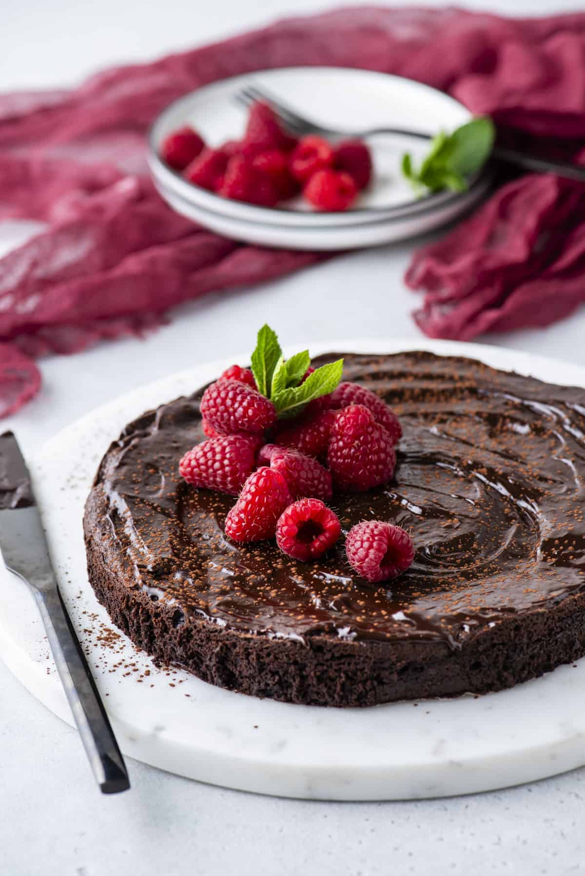 a flourless chocolate cake on a round white serving platter topped with fresh raspberries and mint leaves, with a knife covered in chocolate laying beside it, a red cloth and a bowl of fresh raspberries and mint in the background