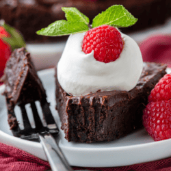 a slice of flourless chocolate cake on a white plate, topped with whipped cream, a fresh raspberry and mint, with a fork laying beside it with one bite on it, more fresh raspberries beside it and more cake in the background
