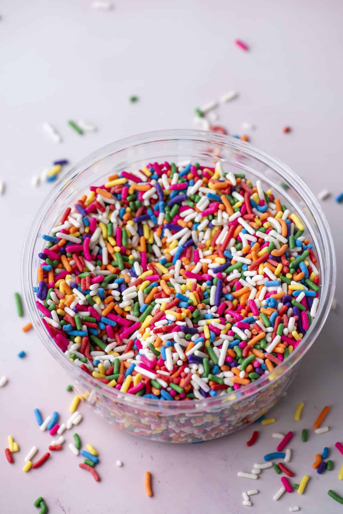 a clear plastic bin of sprinkles with more sprinkles all scattered around it sitting on a light pink surface