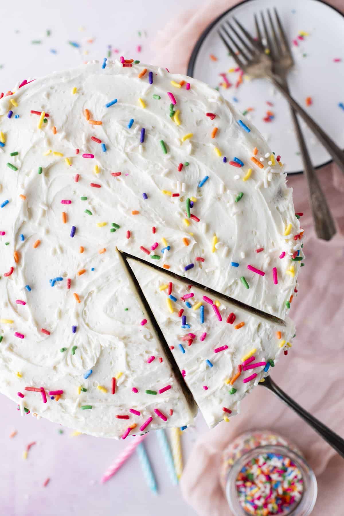 overhead view of one slice of funfetti cake that's been sliced but just barely pulled away from the cake by a spatula, with birthday candles, sprinkles, a plate and forks surrounded the cake stand