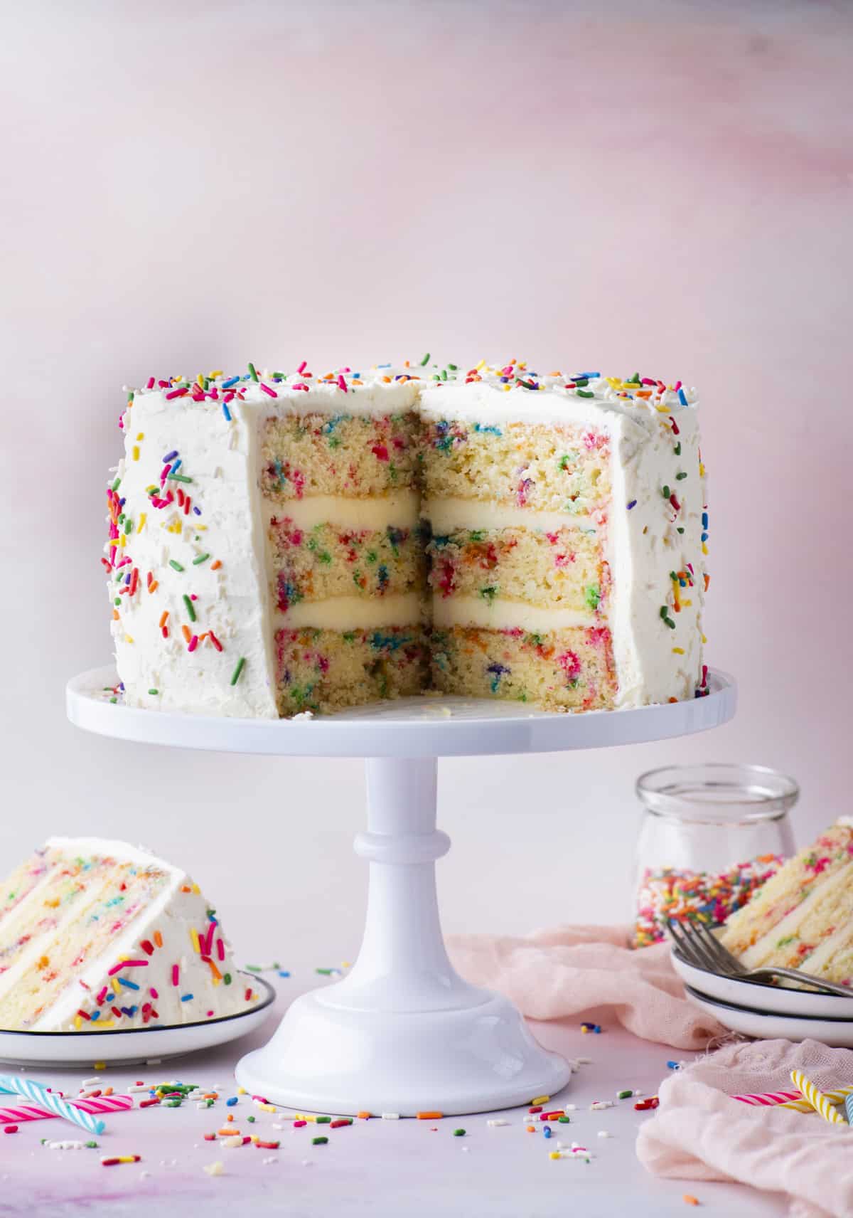 funfetti cake with two slices missing sitting on a white cake stand surrounded by small white plates with the two slices on them, pink towels, birthday candles and scattered sprinkles