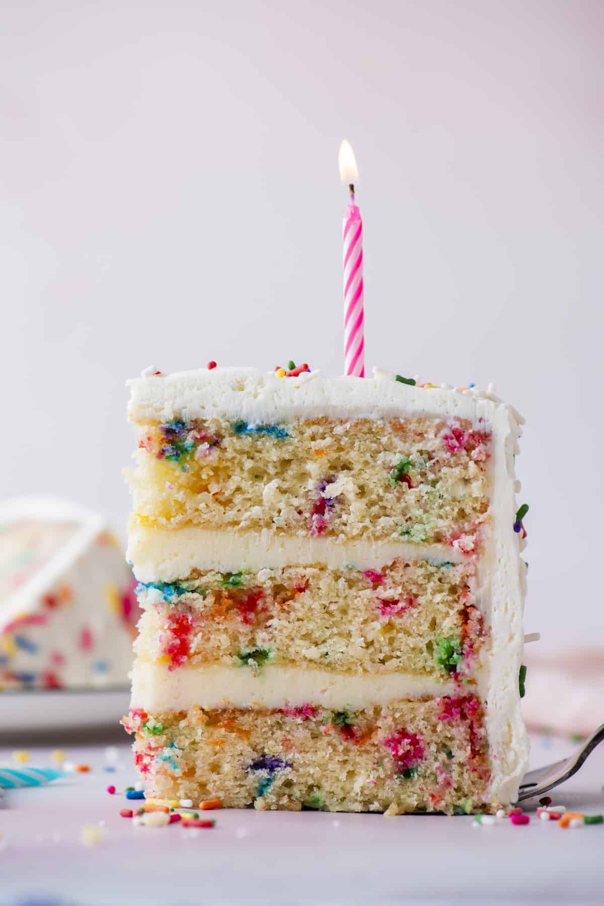a slice of funfetti cake from the side showing its three layers of cake with white icing in between and a lit pink and white striped candle in the top of the slice, with sprinkles scattered around
