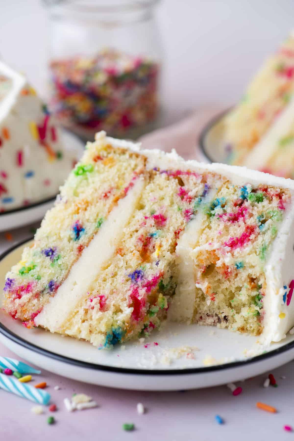 a slice of funfetti cake with a big bite missing sitting on a small white plate surrounded by sprinkles, blue birthday candles and more slices of cake in the background