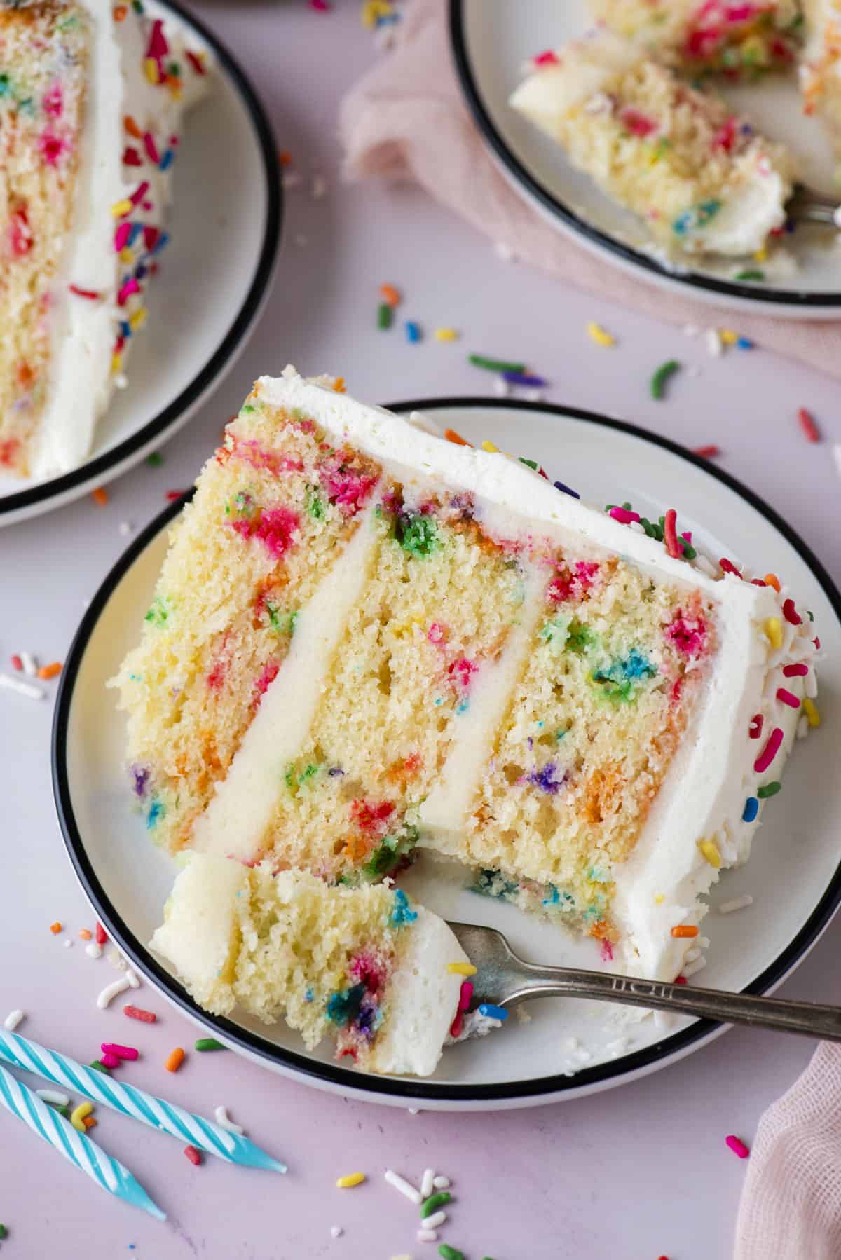 Funfetti cake on three small white plates sitting on a pink surface, with forks that have removed bites of cake from the slice and sprinkles and two blue birthday candles surrounding the plates