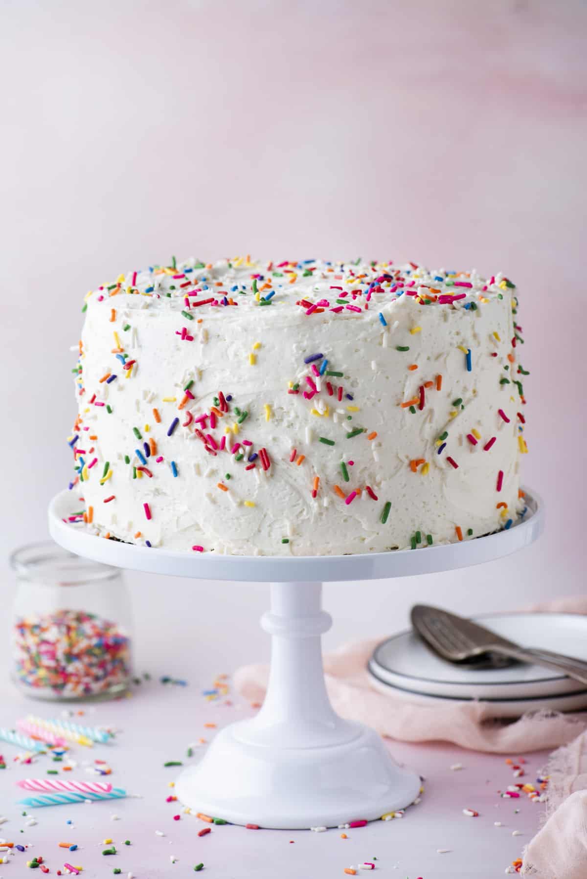 a funfetti cake decorated with white icing and sprinkles on a white cake stand against a pink background with sprinkles, birthday candles, a clear container of sprinkles, plates and utensils surrounding it