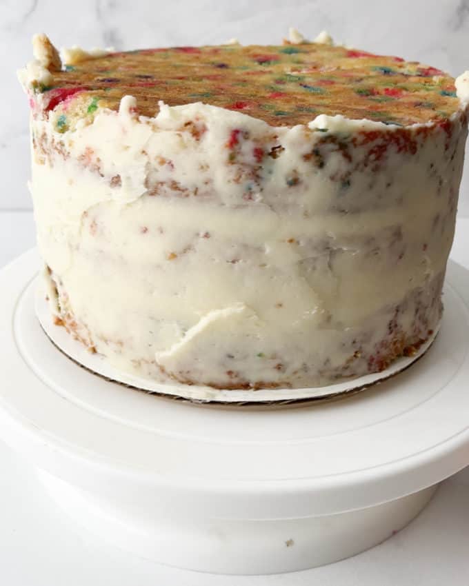 funfetti cake with the sides covered in a thin layer of white icing sitting on top of a white cake stand