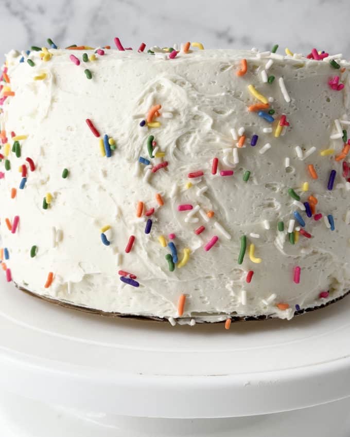 side view of a funfetti cake with white icing and sprinkles covering it sitting on a white cake stand