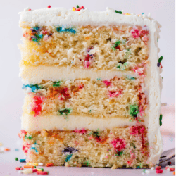 a slice of layered funfetti cake with 3 layers of cake and two layers of frosting, with rainbow sprinkles all around