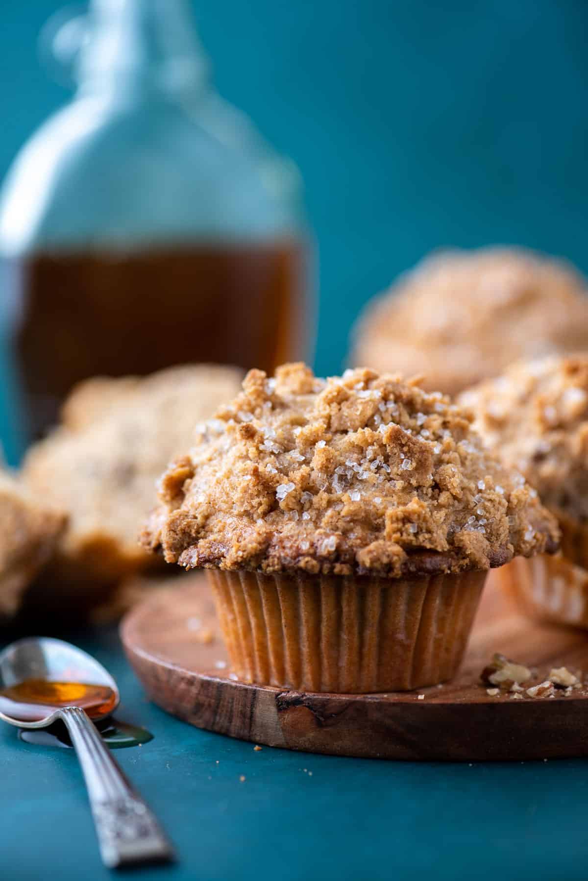 a maple pecan muffin on a wood plate on top of a teal surface with more muffins in the background and a spoon of maple syrup beside it