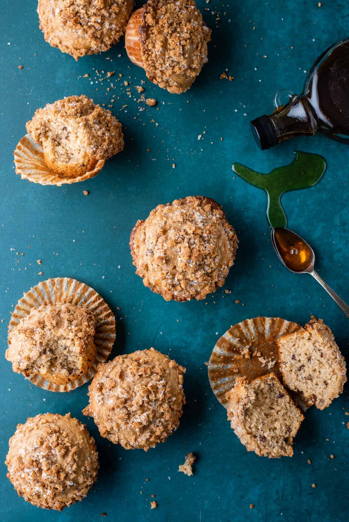 several maple pecan muffins on a dark teal surface, some sitting upright, some on their side, cut in half or with a bite out, with a bottle of maple syrup laying on its side and a spoon with maple syrup on it and drizzled around it