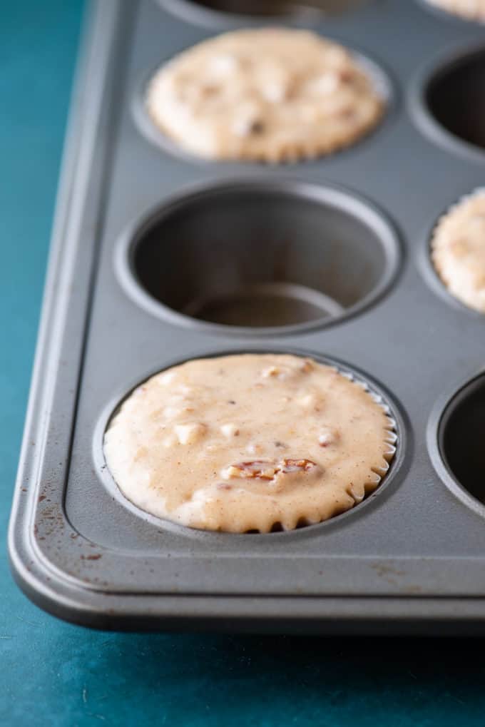 a muffin pan on a dark teal surface with maple pecan muffin batter in every other muffin hole