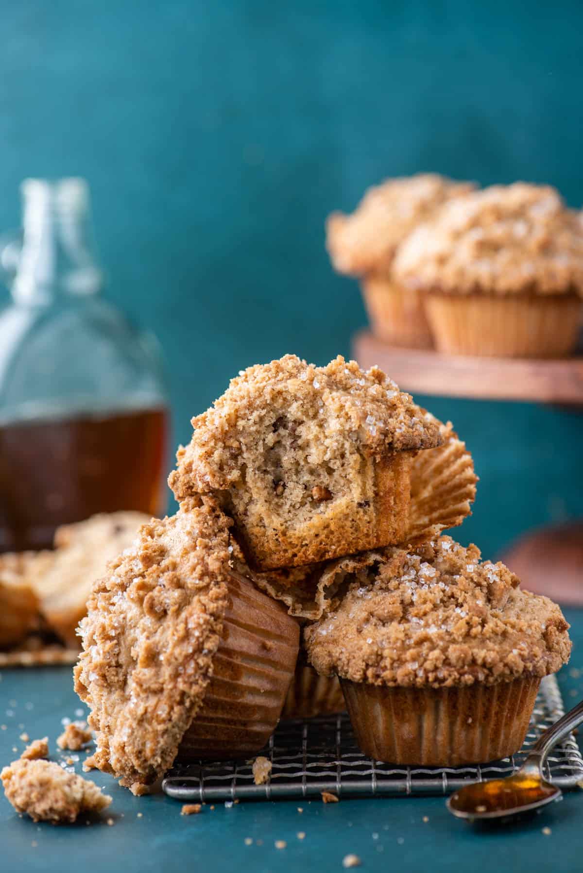 a pile of maple pecan muffins on top of a wire rack, with the top muffin missing a bite out, a spoon full of maple syrup, and more muffins and maple syrup in the background
