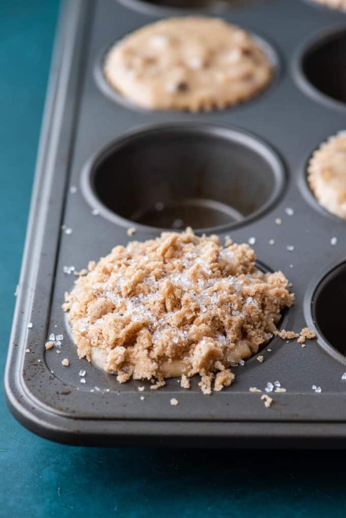 a muffin pan on a dark teal surface with maple pecan muffins in every other muffin hole topped with crumb topping and sanding sugar