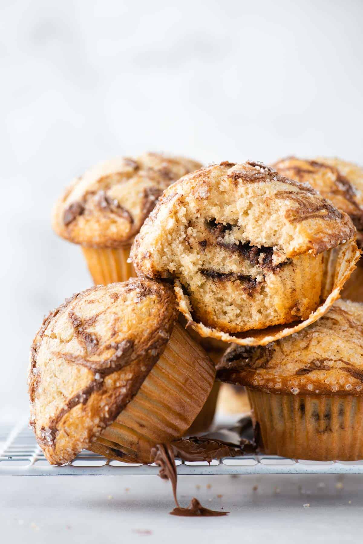 a pile of nutella swirl muffins on a wire rack, with one muffin on its side and the one on top of it with the muffin paper pulled partially off, exposing the layers of nutella inside, and a knife with nutella dripping down from the wire rack