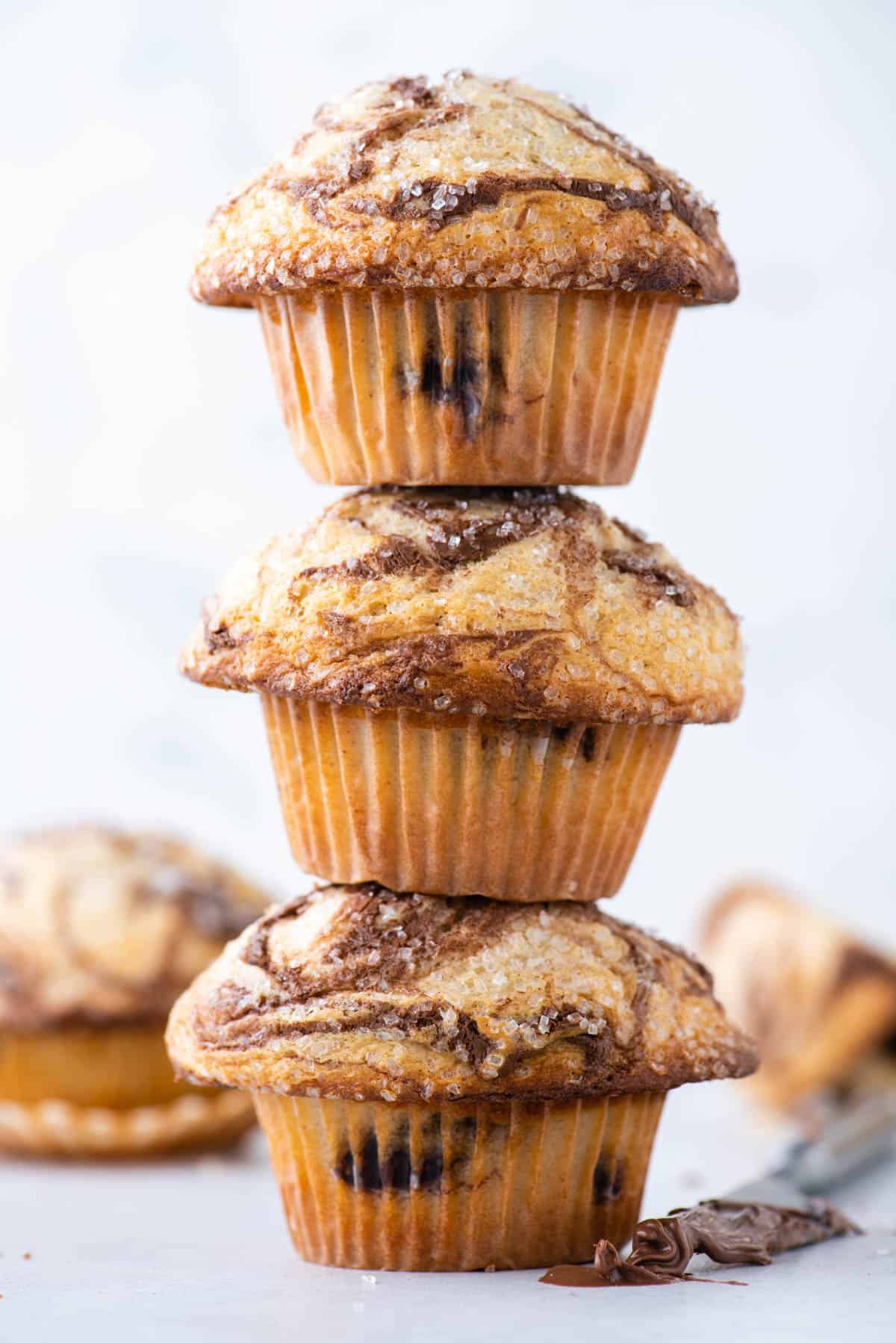 a tower of 3 nutella swirl muffins with more muffins in the background and a butter knife with nutella on it beside the stack