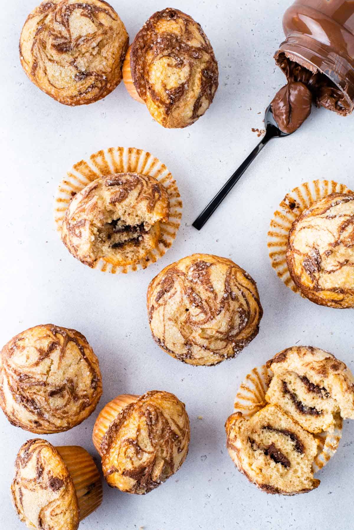 several nutella swirl muffins arranged on a white surface, with some muffins sitting upright, some on their sides, one cut in half exposing the center, and one with a bite out of it, with a nutella jar laying on its side and a spoon of nutella laying beside its opening