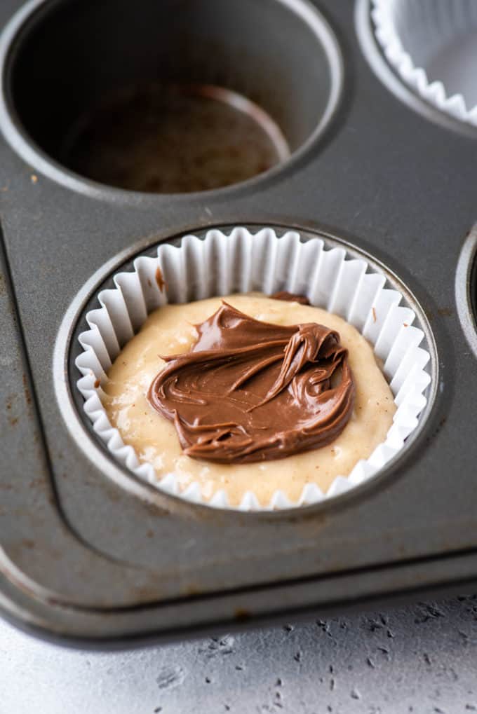 a muffin pan with a white muffin liner in the corner spot and some muffin batter in the bottom of it and layers of muffin batter and nutella inside the liner