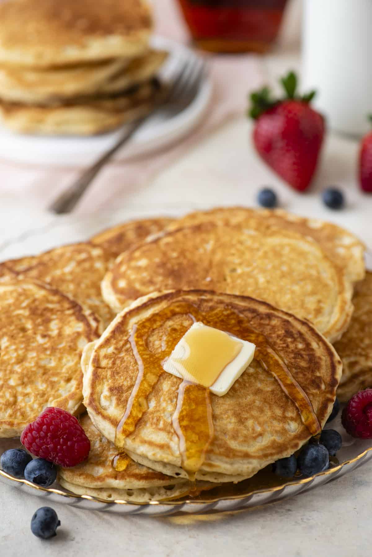 pancakes piled on a plate with one topped with butter and drizzled with syrup, with fresh berries surrounding them and more berries and pancakes in the background
