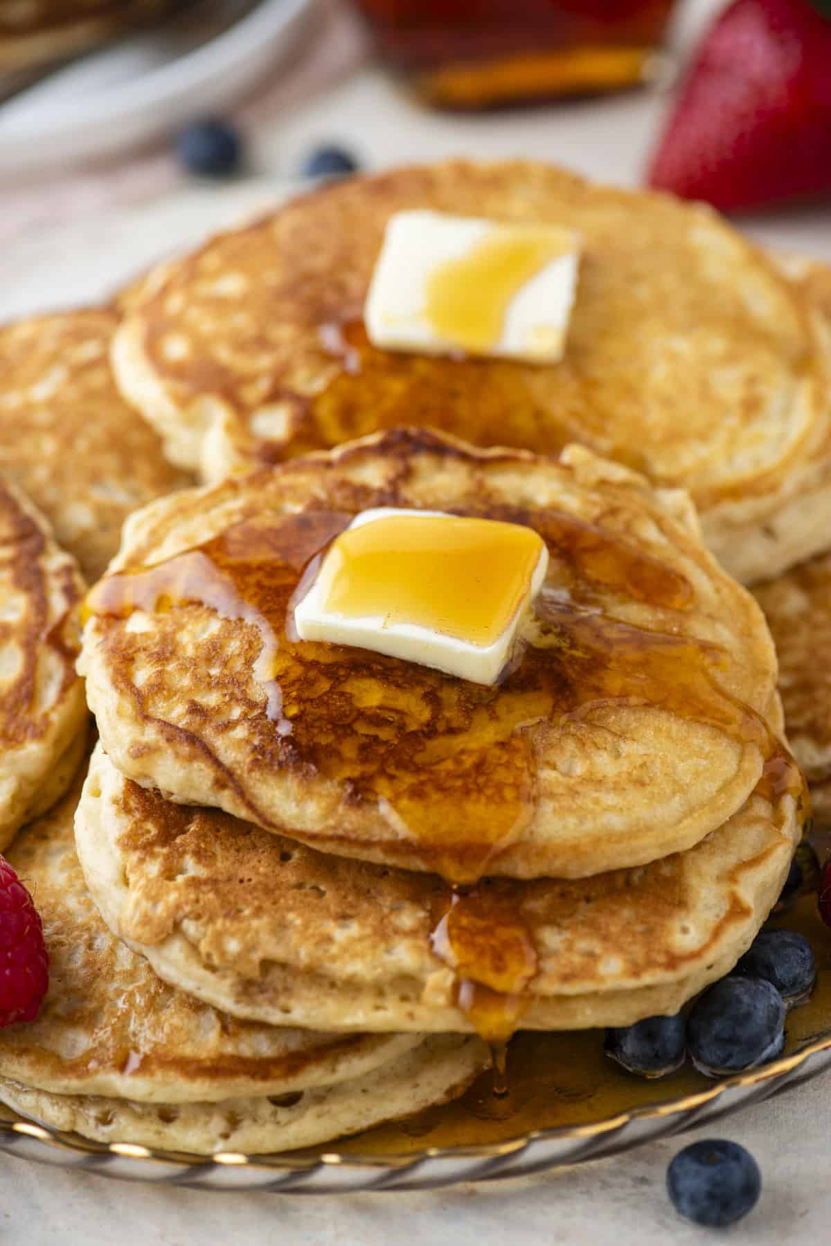 pancakes piled on a plate topped with butter and drizzled with syrup, with fresh berries surrounding them and more berries and pancakes in the background